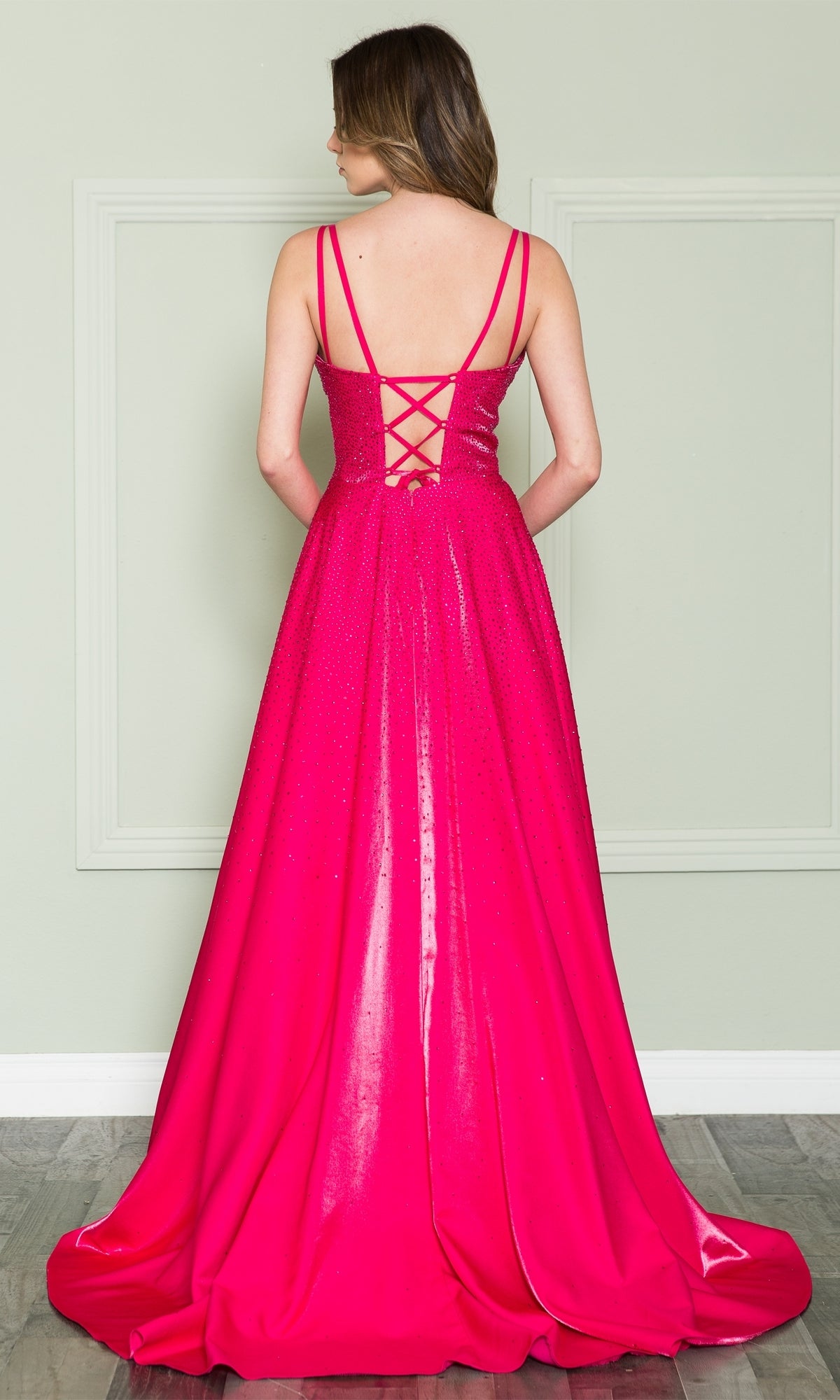 A woman stands facing away, wearing the Poly USA 8922 long beaded a-line prom dress in hot pink with thin straps and a lace-up back, posed against a light-colored wall with molding.